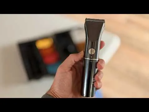 A hand holds the sleek, black The Beard Club Beard Trimmer in a clear close-up, with the case of colorful guide combs blurred in the background.
